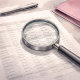 Editorial illustration of a magnifying glass examining rows of figures on a financial document, with a magenta leaflet pushed aside on a desk.