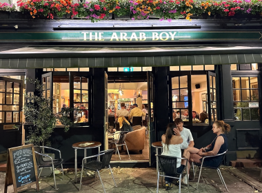 Open-front pub named The Arab Boy with colorful flowers above, outdoor seating and patrons inside and outside.