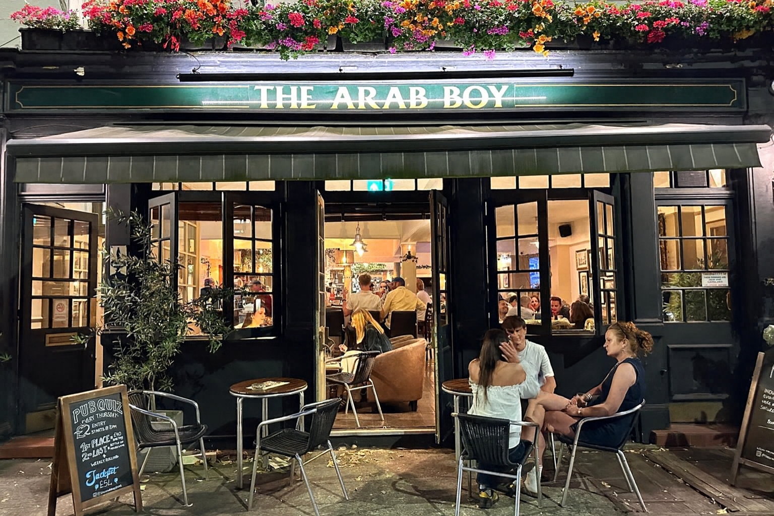 Open-front pub named The Arab Boy with colorful flowers above, outdoor seating and patrons inside and outside.