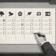 Overhead editorial illustration of a monochrome UK local election ballot paper on a grey polling booth shelf, with five party-style icons across the top, two boxes marked with black Xs, and a hand holding a pen hovering over an empty box.