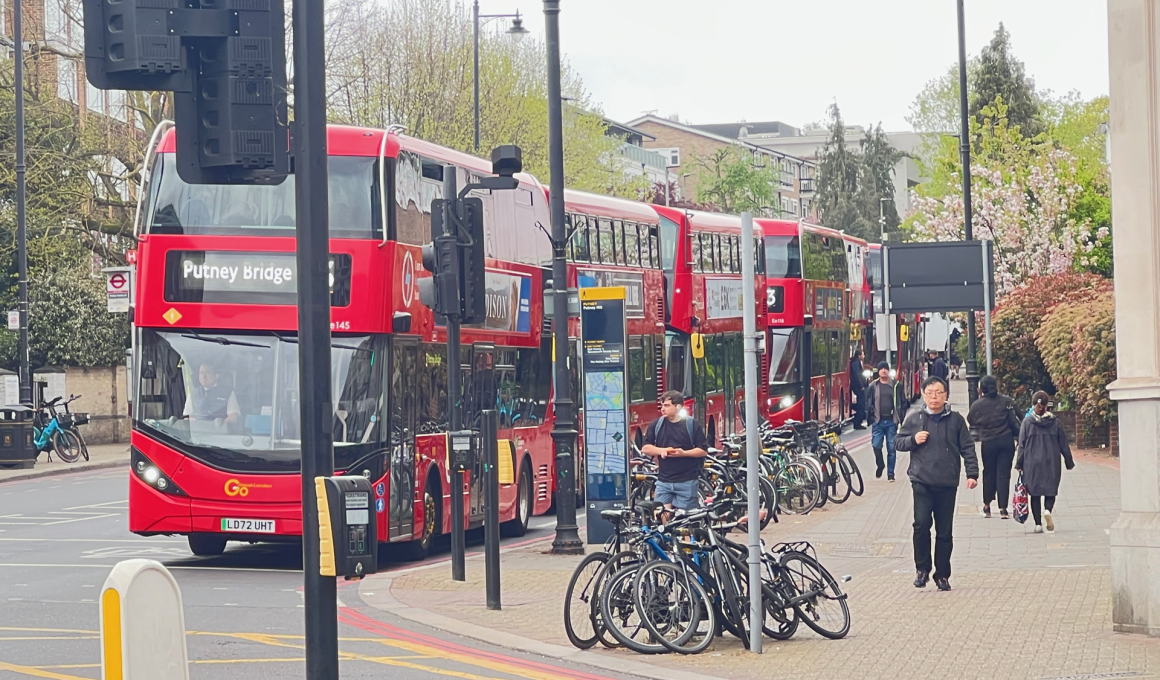 Red double-decker bus with a 'Putney Bridge' destination sign, parked on a busy city street beside a bike rack and pedestrians nearby.