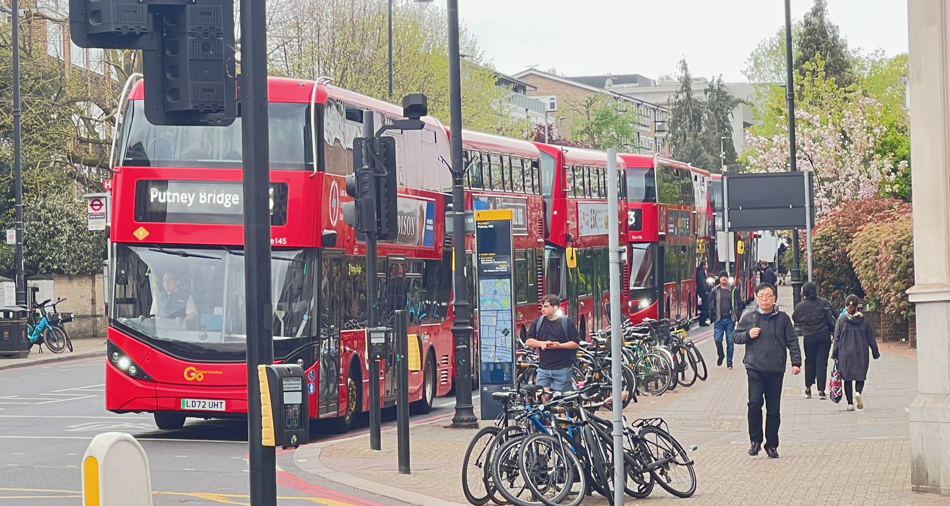Red double-decker bus with a 'Putney Bridge' destination sign, parked on a busy city street beside a bike rack and pedestrians nearby.