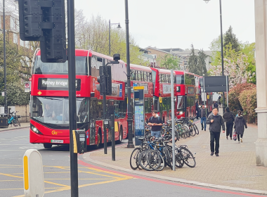 Red double-decker bus with a 'Putney Bridge' destination sign, parked on a busy city street beside a bike rack and pedestrians nearby.