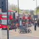 Red double-decker bus with a 'Putney Bridge' destination sign, parked on a busy city street beside a bike rack and pedestrians nearby.