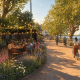 Outdoor riverside cafe with diners under string lights, bordered by colorful flowers and trees at golden hour beside a calm river.