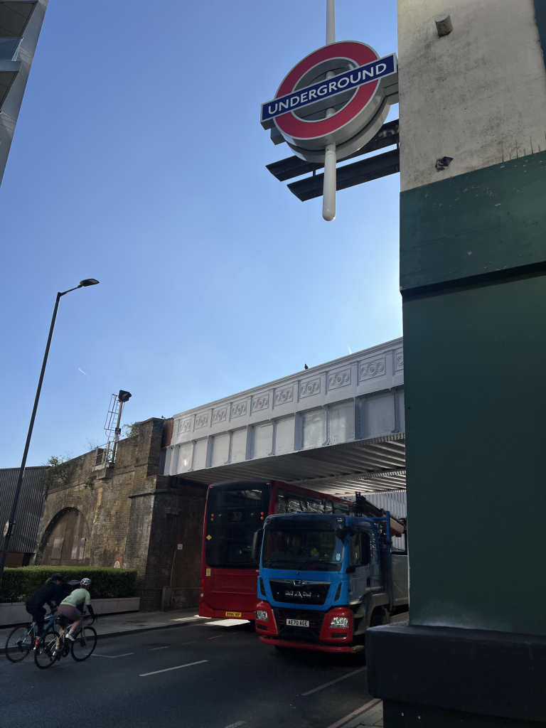 London Underground roundel sign mounted on a pillar above a red and blue bus on a city street.