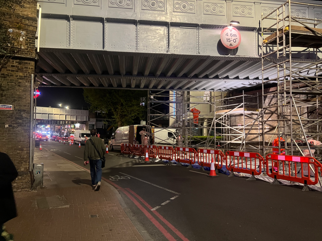 Nighttime construction under a steel bridge with scaffolding, orange barriers, and workers in safety gear.