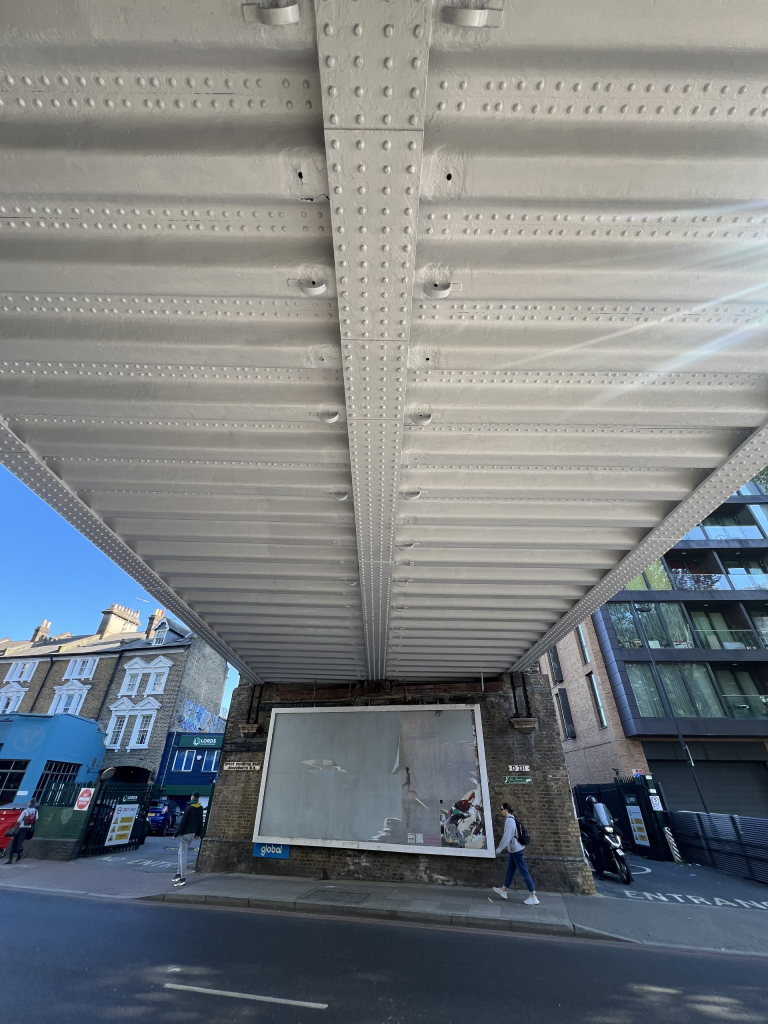Underside of a riveted metal bridge spanning a city street, with pedestrians and buildings visible beneath.
