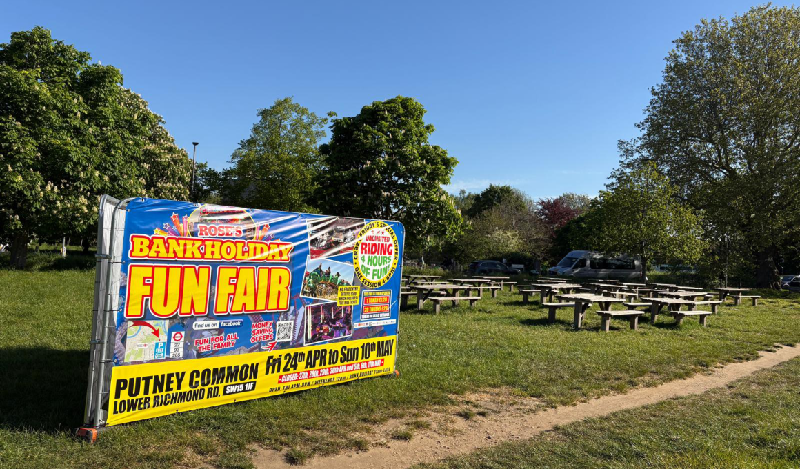 Colorful banner reading 'BANK HOLIDAY FUN FAIR' advertising an event, set up beside a grassy park with picnic tables nearby.