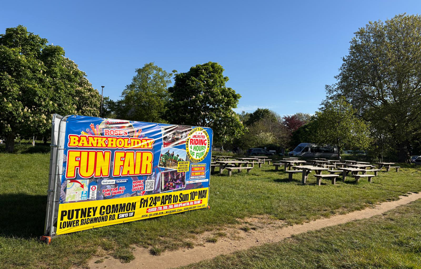 Colorful banner reading 'BANK HOLIDAY FUN FAIR' advertising an event, set up beside a grassy park with picnic tables nearby.