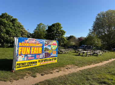 Colorful banner reading 'BANK HOLIDAY FUN FAIR' advertising an event, set up beside a grassy park with picnic tables nearby.