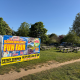 Colorful banner reading 'BANK HOLIDAY FUN FAIR' advertising an event, set up beside a grassy park with picnic tables nearby.