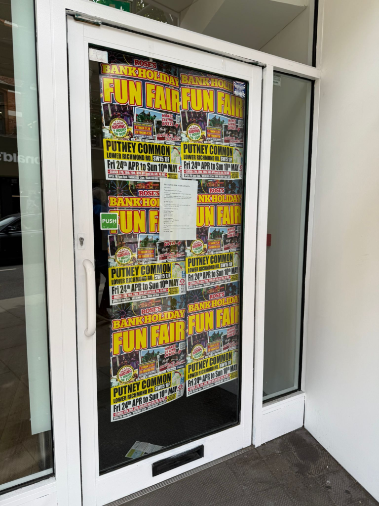 Glass door covered with bright, busy posters advertising a bank holiday fun fair at Putney Common on Lower Richmond Rd, with large 'FUN FAIR' text.