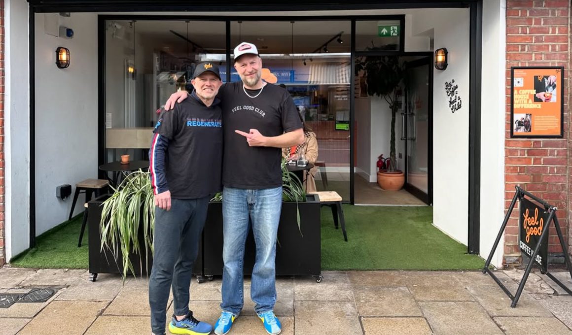 Two men stand outside a cafe with their arms around each other, smiling at the camera, in front of glass doors, potted plants and outdoor seating.