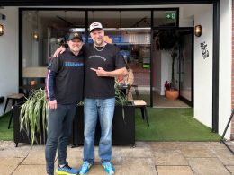 Two men stand outside a cafe with their arms around each other, smiling at the camera, in front of glass doors, potted plants and outdoor seating.