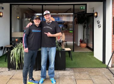 Two men stand outside a cafe with their arms around each other, smiling at the camera, in front of glass doors, potted plants and outdoor seating.