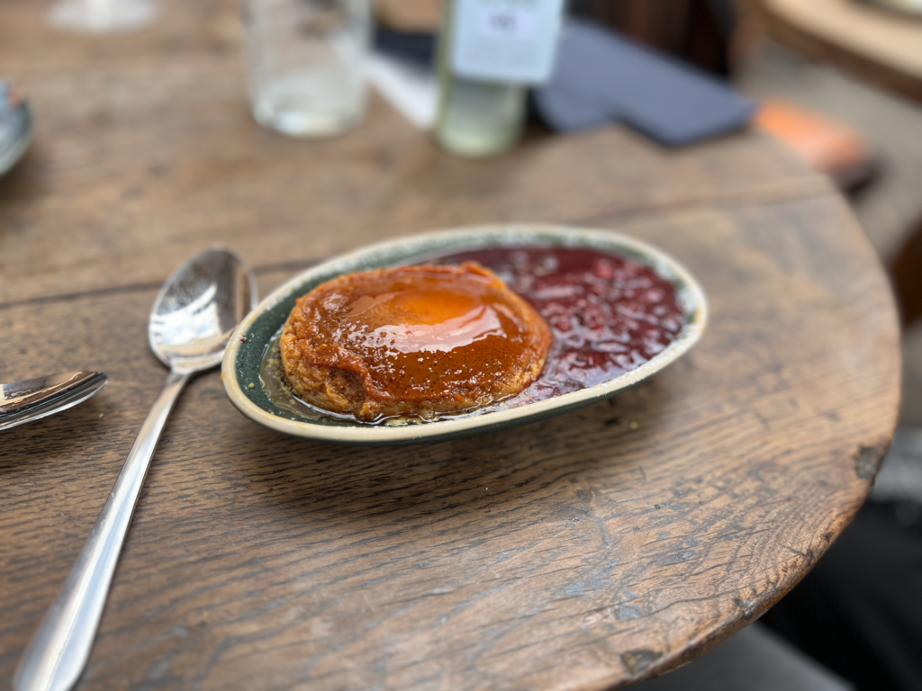 Caramel flan on a green-rimmed plate with red sauce on a rustic wooden table, with a spoon and fork nearby