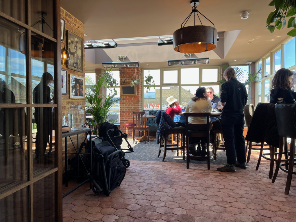 Sunlit café interior with brick walls, framed art, and plants; a group sits around a tall table by large windows, chatting.