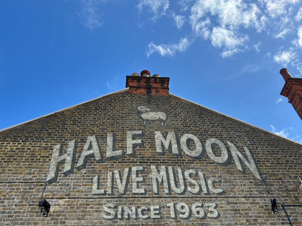 Faded white sign on a brick wall reading 'HALF MOON LIVE MUSIC SINCE 1963' with a small sheep graphic, under a bright blue sky.