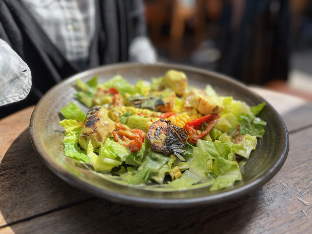 Plate of grilled vegetable salad with lettuce, corn, zucchini and red peppers on a dark ceramic dish outdoors on a wooden table.