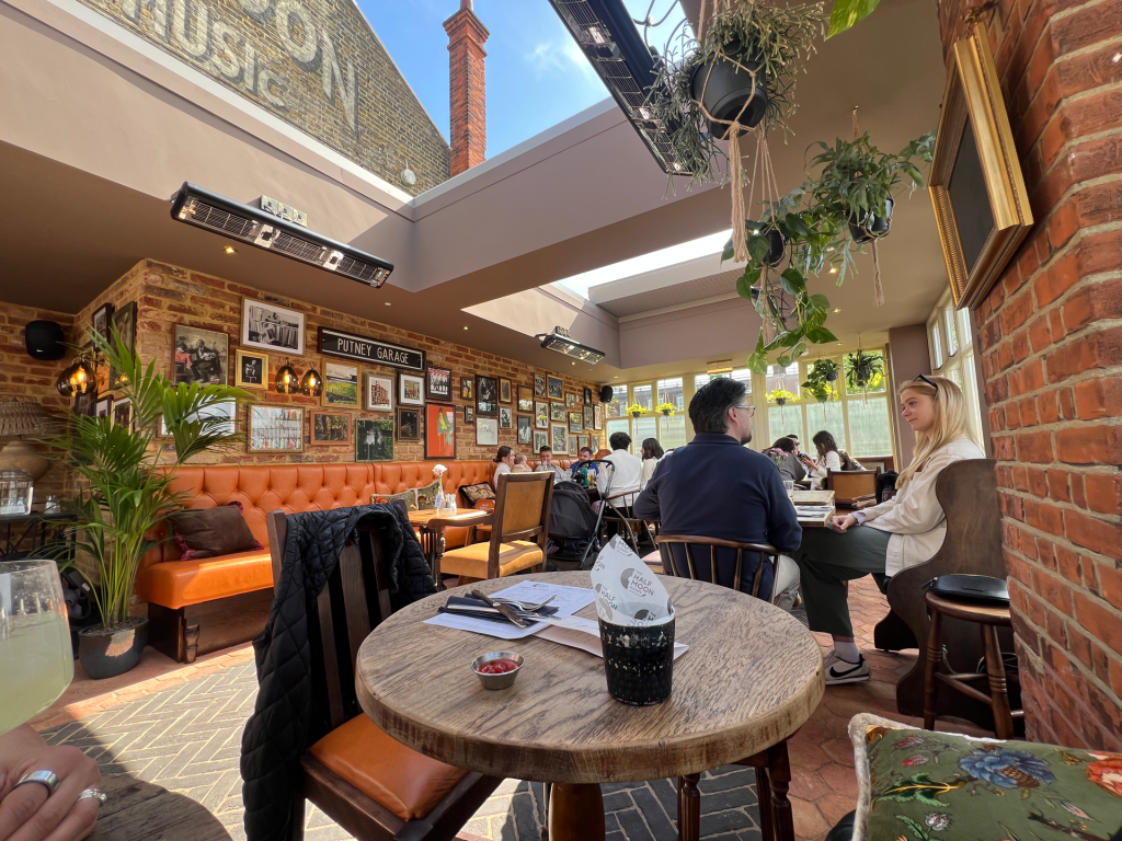 Bright cafe interior with exposed brick, an orange tufted banquette, wall photo gallery, hanging plants, and customers dining by a skylight.