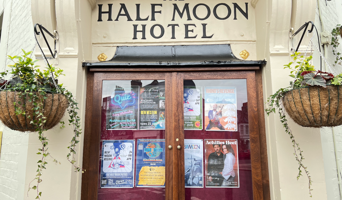 Framed wooden double doors of 'The Half Moon Hotel' with posters displayed in glass panels and two hanging planters on either side.