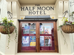 Framed wooden double doors of 'The Half Moon Hotel' with posters displayed in glass panels and two hanging planters on either side.