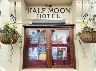 Framed wooden double doors of 'The Half Moon Hotel' with posters displayed in glass panels and two hanging planters on either side.