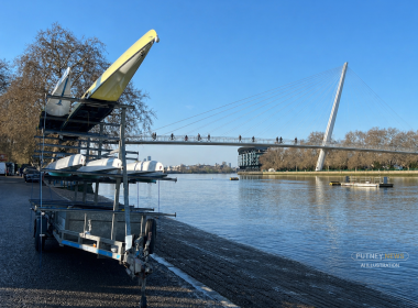 Trailer loaded with kayaks on the riverbank beside a cable-stayed pedestrian and bike bridge over a calm river, with bare trees in the background.
