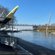 Trailer loaded with kayaks on the riverbank beside a cable-stayed pedestrian and bike bridge over a calm river, with bare trees in the background.