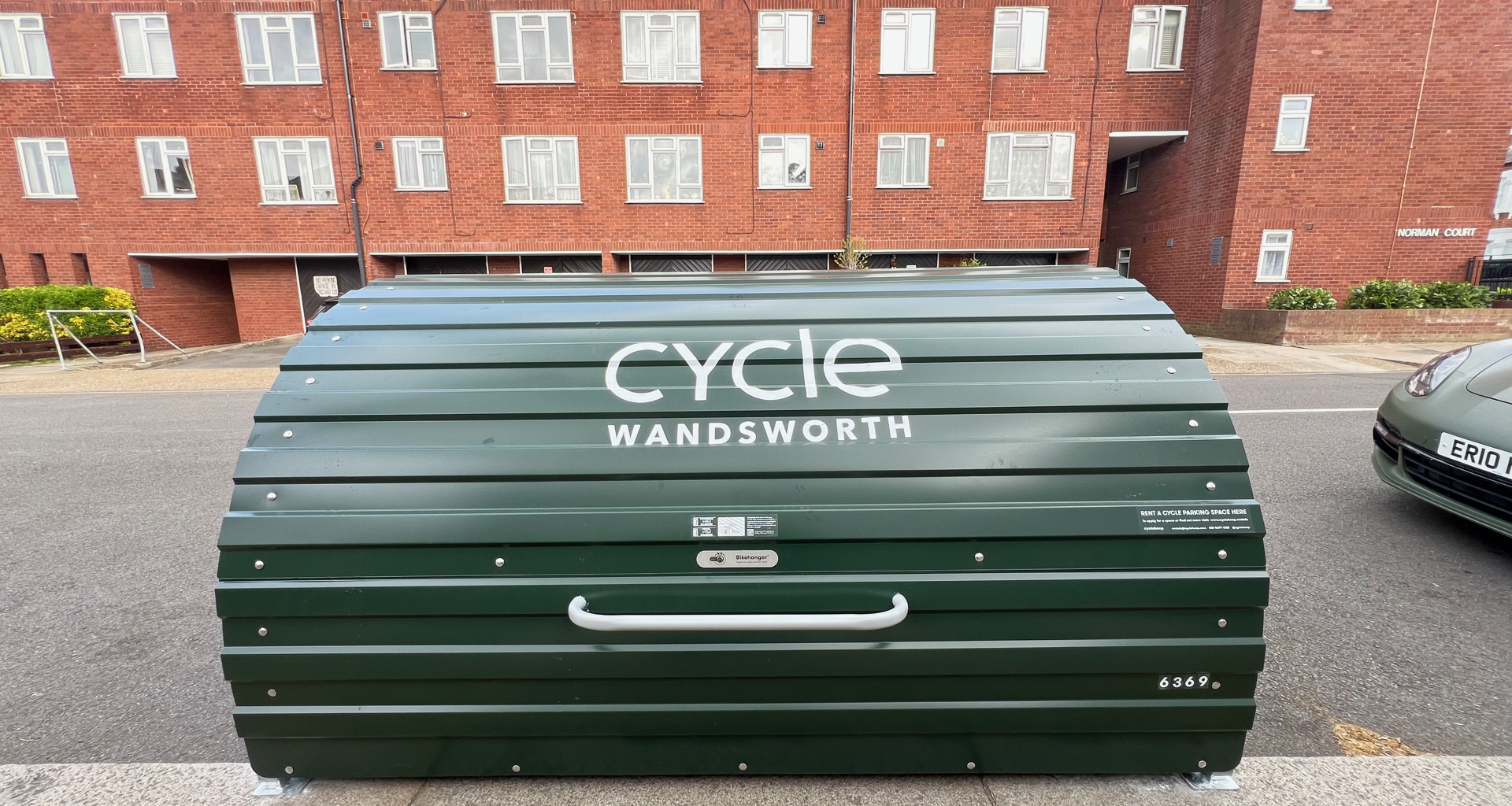 Green curved cycle storage unit with 'cycle Wandsworth' lettering parked on a street in front of red brick apartment buildings