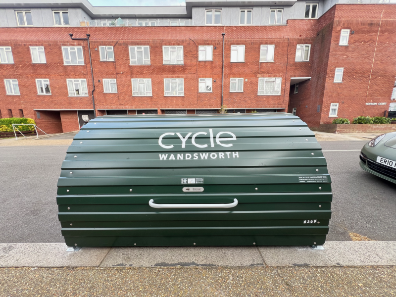 Green curved cycle storage unit with 'cycle Wandsworth' lettering parked on a street in front of red brick apartment buildings