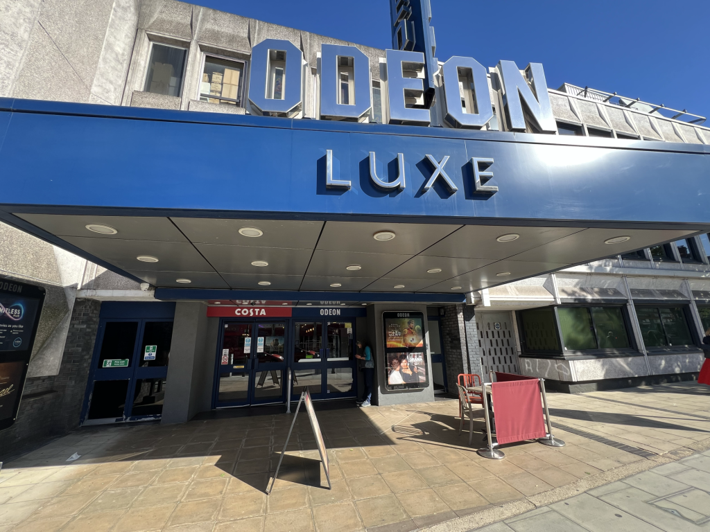 Front of an Odeon Luxe cinema with large silver letters on a blue marquee and glass doors beneath. Posters visible by the entrance.