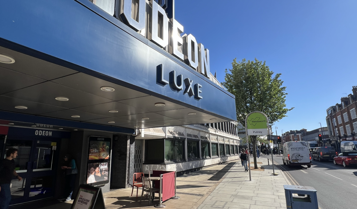 Blue Odeon Luxe cinema façade with large 3D letters along a sunny street and pedestrians nearby.