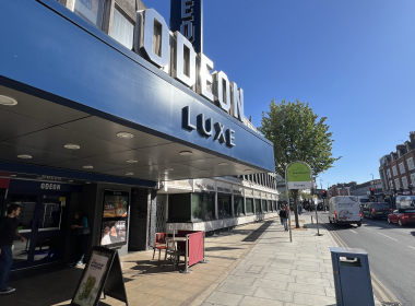 Blue Odeon Luxe cinema façade with large 3D letters along a sunny street and pedestrians nearby.