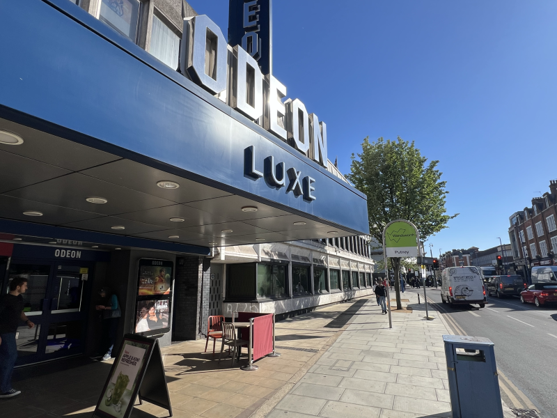 Blue Odeon Luxe cinema façade with large 3D letters along a sunny street and pedestrians nearby.