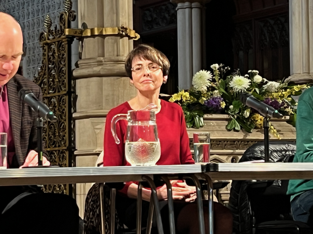 roehampton-hustings - 3 - Putney.news Panel discussion scene with a woman in a red top seated at a long table, microphones in front and a water pitcher nearby, ornate backdrop and flowers behind.