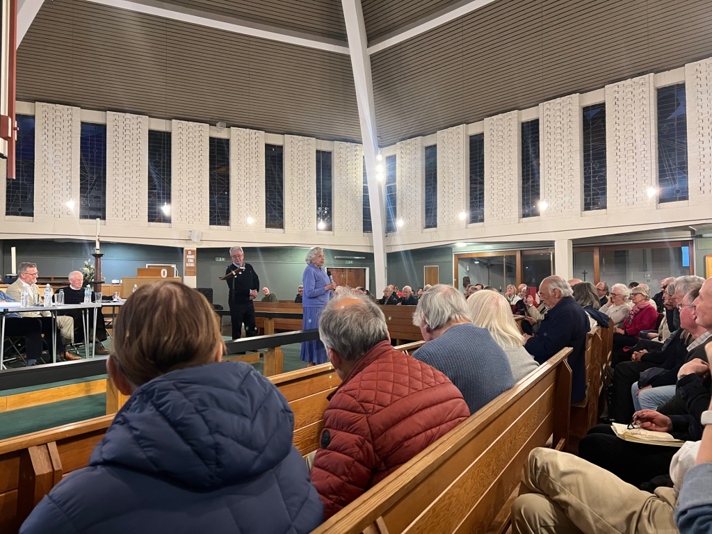 Crowded church hall with attendees seated on wooden pews while a woman in a blue dress speaks at the front near a lectern.