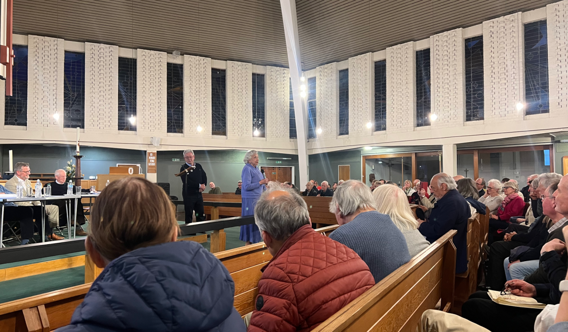Crowded church hall with attendees seated on wooden pews while a woman in a blue dress speaks at the front near a lectern.
