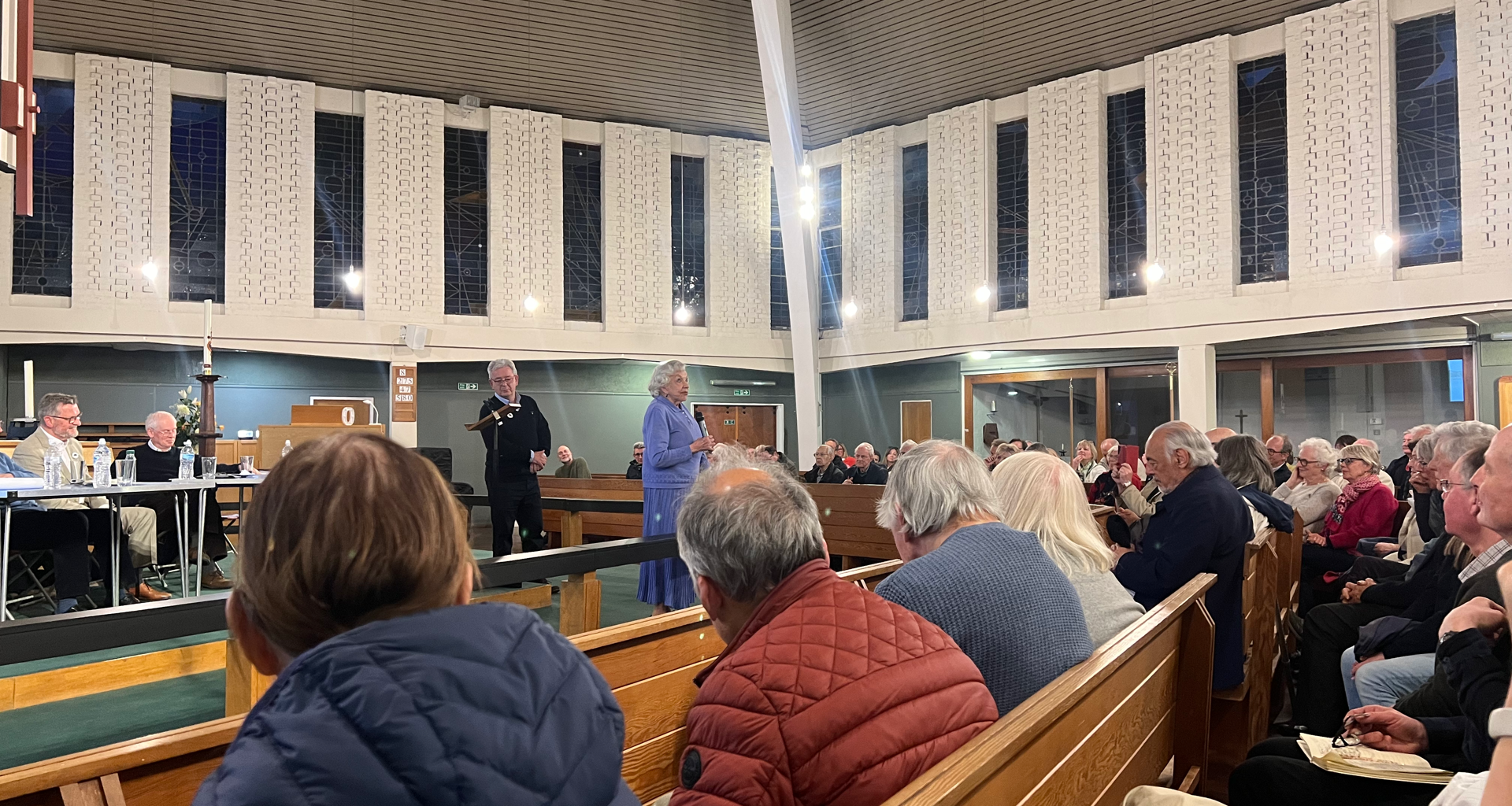 Crowded church hall with attendees seated on wooden pews while a woman in a blue dress speaks at the front near a lectern.