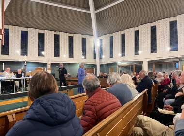 Crowded church hall with attendees seated on wooden pews while a woman in a blue dress speaks at the front near a lectern.