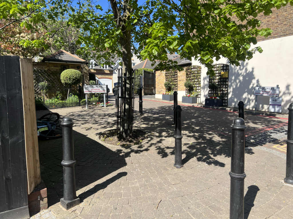 A tree-lined cobblestone courtyard with planters and gated entrances, signs reading 'Angel Mews' indicating private access.