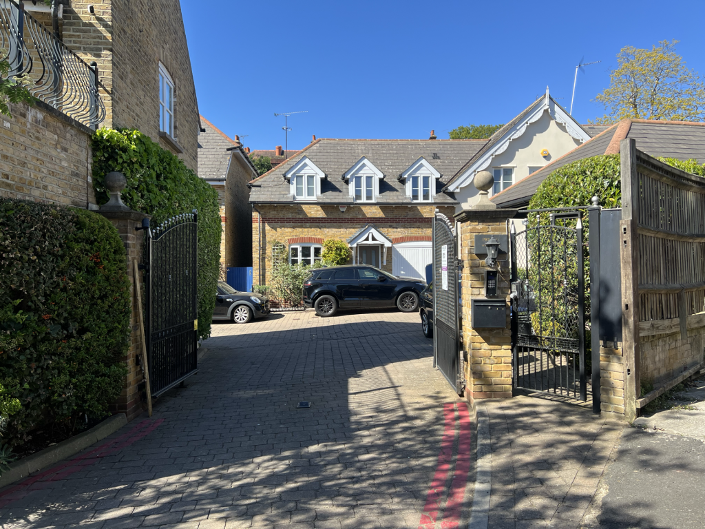 A gated residential driveway with brick houses, parked cars, and a bright blue sky above.