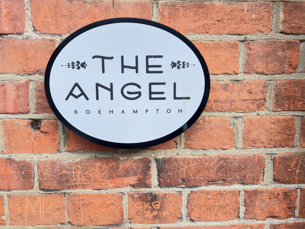 Round white oval sign reading 'THE ANGEL ROEHAMPTON' mounted on a red brick wall.