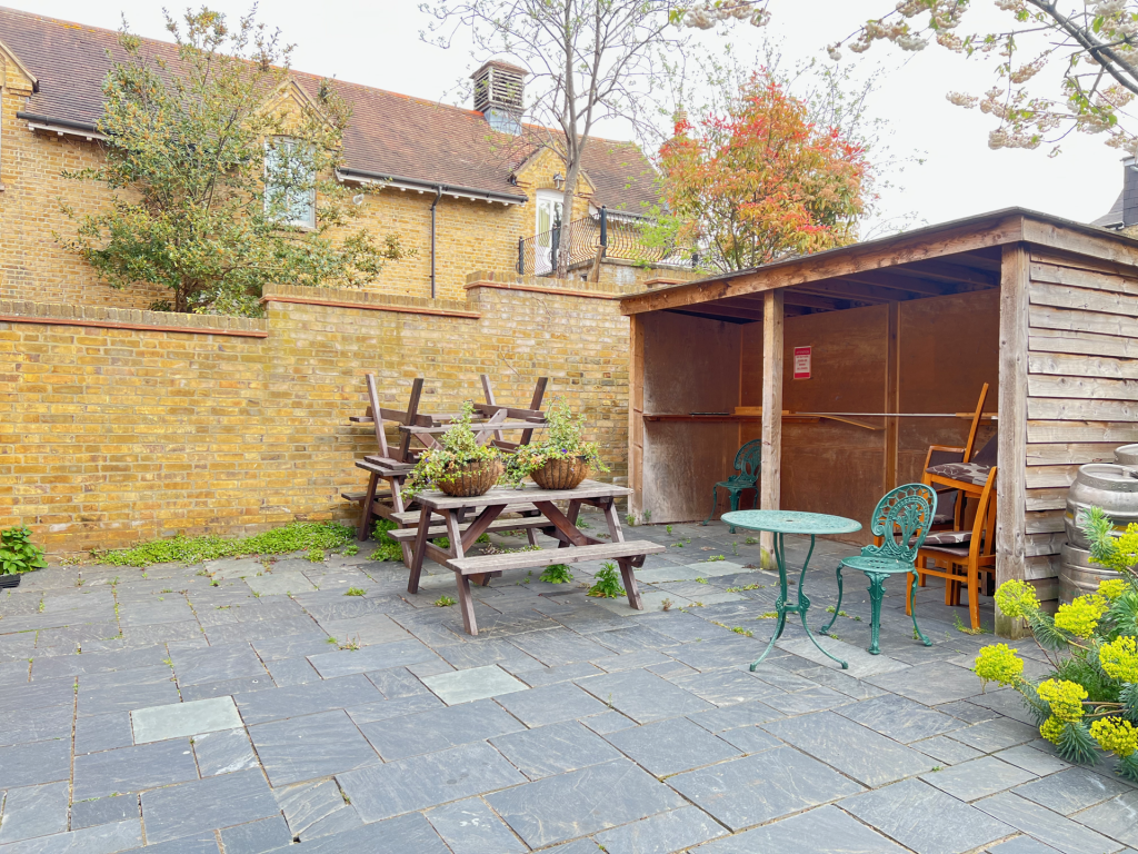 A small backyard patio with a wooden shed, pastel-green metal chairs, a round teal metal table, and stacked wooden benches along a brick wall. Potted plants sit on the tables and yellow flowers bloom nearby.