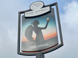 Signboard for The Angel pub showing a silhouette of a woman raising a glass against a sunset, with EST. 1831 at the top