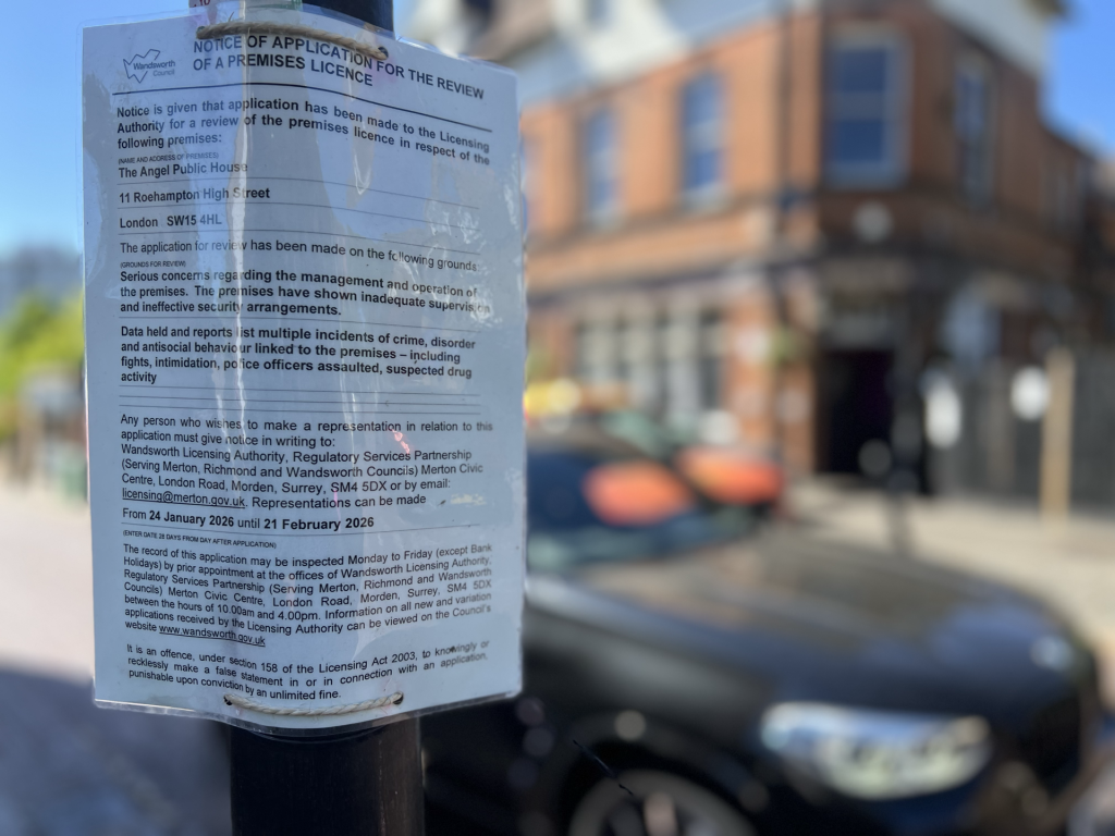 Public notice poster attached to a street pole announcing a license review for a local premises, with a blurred city street in the background and brick buildings nearby.