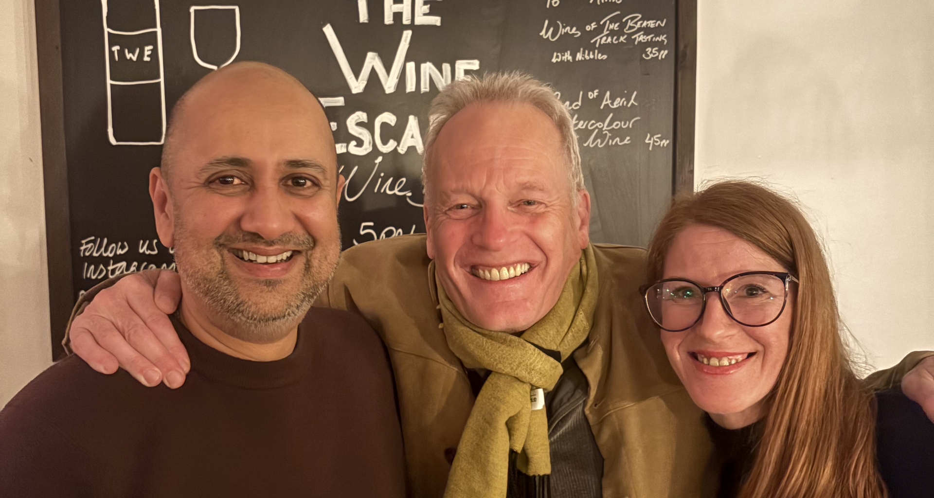 Three smiling adults pose for a selfie in front of a chalkboard menu with wine illustrations in a cozy setting.