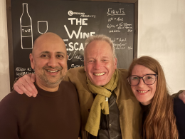 Three smiling adults pose for a selfie in front of a chalkboard menu with wine illustrations in a cozy setting.