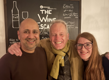 Three smiling adults pose for a selfie in front of a chalkboard menu with wine illustrations in a cozy setting.